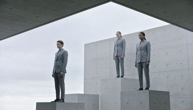 Standing trio wearing grey suits on concrete pedestals in minimalist geometric courtyard