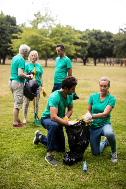 Group of volunteers wearing green shirts collecting litter in a park. They are using garbage bags and gloves to pick up trash, demonstrating teamwork and community spirit. Ideal for promoting environmental awareness, community events, and social responsibility initiatives.