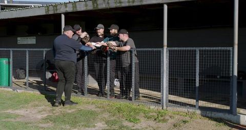 Sunlit team huddle passing papers over mesh fence at dugout during casual baseball meeting