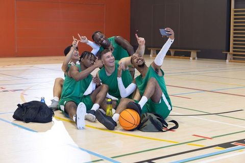 Diverse male basketball team celebrating and taking selfie while resting on indoor court