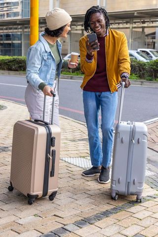 Smiling travelers checking phone while waiting with luggage on city curb