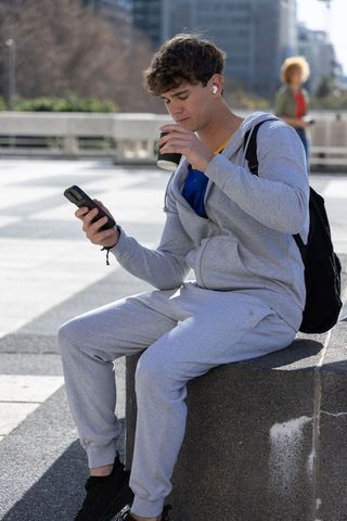 Teen male in grey sweats checking smartphone while sipping coffee on urban plaza bench