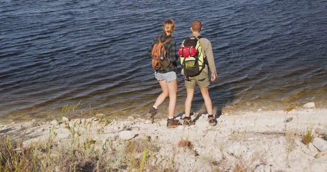 Couple of hikers with backpacks resting by shore of a serene lake. Perfect for travel blogs, outdoor activity promotions, adventure gear advertisements, or articles on summer recreational activities.