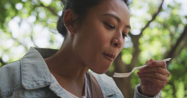 Young Asian woman enjoying picnic food in a park, savoring a bite. Suitable for use in projects related to lifestyle, outdoor activities, food enjoyment, leisure time, and female individual experiences.