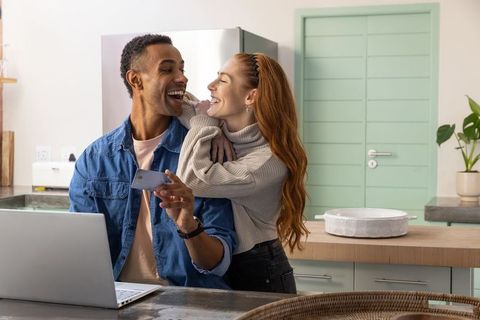 Diverse Couple Laughing in Modern Kitchen with Smartphone and Laptop