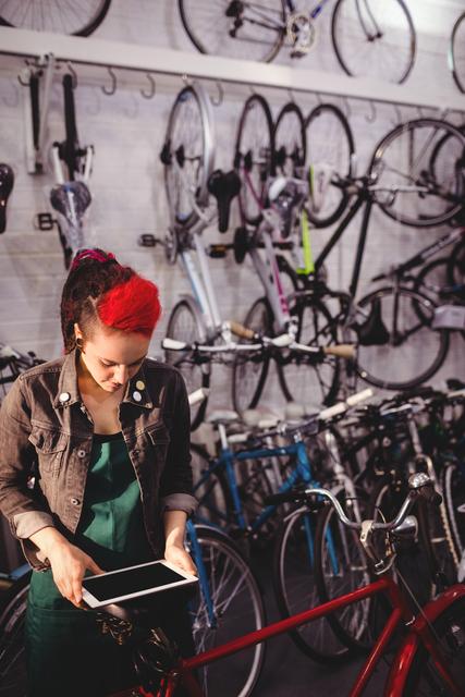 Female bike mechanic with red hair using tablet in a bicycle workshop. Surrounded by bikes and repair tools, she is focused on her device, possibly checking information or ordering parts. Suitable for use in themes related to modern technology in traditional professions, female professionals in technical fields, or advertisements for bike shops and repair services.