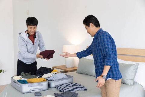 Asian male friends packing suitcase together in bright bedroom preparing for trip