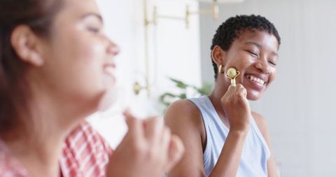 Diverse female friends enjoying skincare routine using gold facial roller and moisturizer