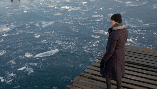 Solitary woman standing on frozen pier looking over icy water in fur-trim coat and beanie