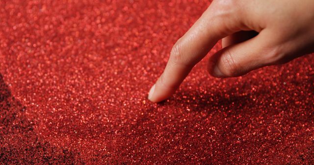 Close-up of a hand touching a red glitter surface. Ideal for use in crafting, decoration projects, holiday themes, artistic visuals, and design textures. Emphasizes tactile engagement with shiny materials. Suitable for websites, presentations, and marketing materials requiring a touch of sparkle.