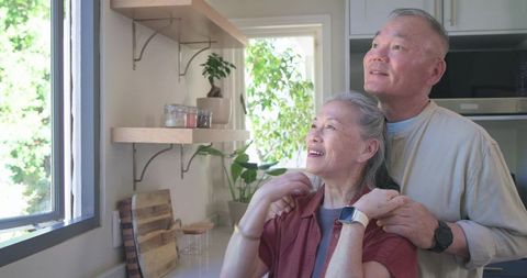 Senior asian couple enjoying morning light in cozy kitchen with smartwatches and greenery