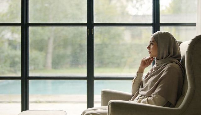Contemplative woman wearing hijab gazing through large paned window over calm pool serene interior