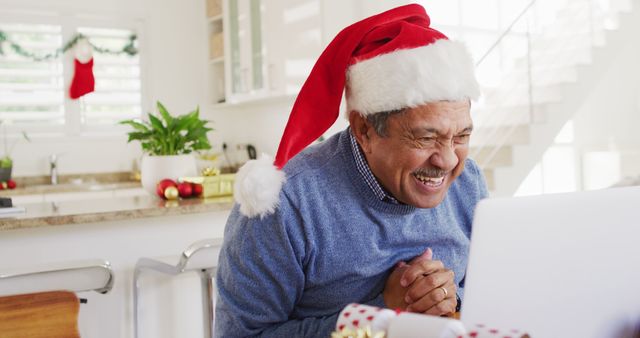 Image of happy senior biracial man in santa hat making christmas laptop image call, holding gift. Christmas, tradition, global communication, inclusivity and senior lifestyle concept.