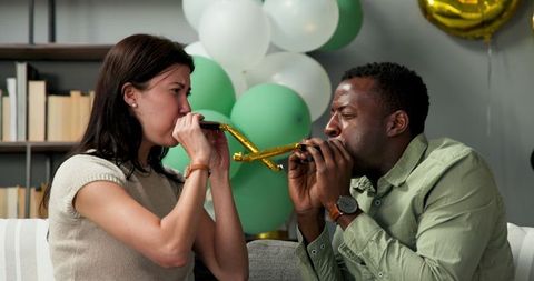 Joyful couple blowing birthday party horns on sofa