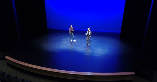 Two performers, actors, stand on a stage with a vibrant blue backdrop, engaging in a scene or dialogue. Their presence on stage suggests a theatrical performance or rehearsal in progress, capturing a moment of creative expression.