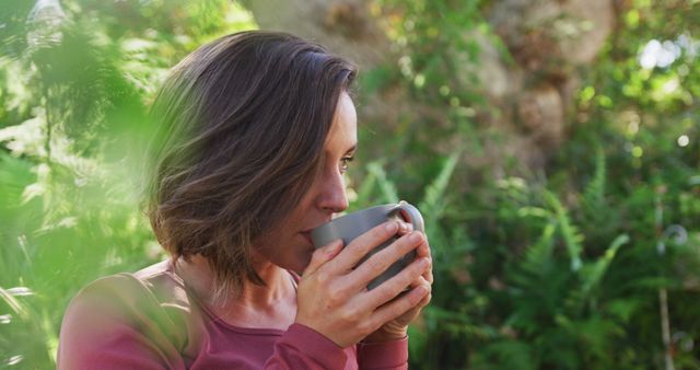 Woman sipping coffee surrounded by lush green foliage, creating a peaceful and relaxing atmosphere. Perfect for themes of relaxation, enjoying nature, outdoor activities, morning routines, and tranquility.