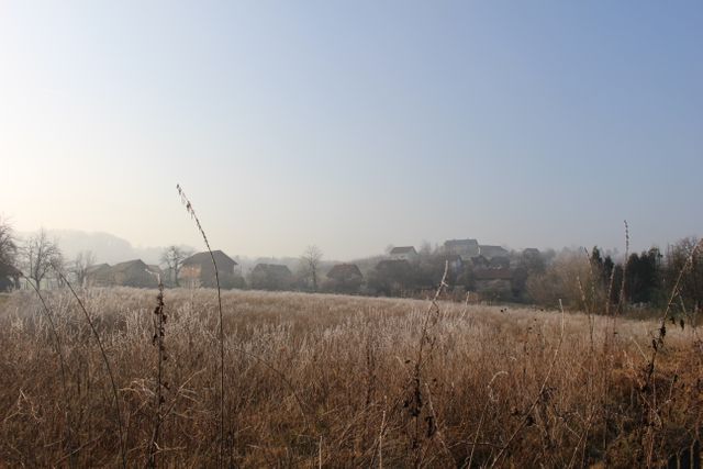 This stock photo depicts a foggy morning in a rural countryside with frosty fields and a small village in the background. Ideal for illustrating serene rural life, weather conditions, nature photography, travel brochures, or agricultural themes. Could be used in blogs, social media posts, or printed materials related to tranquility, relaxation, and natural beauty.