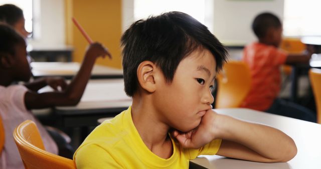 Bored asian schoolboy sitting at desk in diverse elementary school class. Childhood, education, learning and elementary school, unaltered.