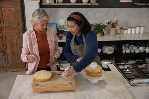 Senior friends frosting layered cake together in cozy home kitchen, baking and sharing
