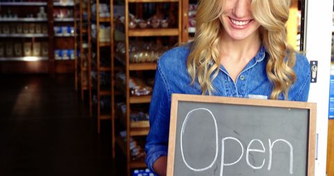 Smiling Female Shop Owner Holding Open Sign at Grocery Store Entrance