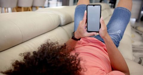 a young man is lounging comfortably on a cream-colored sofa. he has curly hair and is wearing a casual pink t-shirt with denim shorts. the focus is on his arms as he holds a smartphone, featuring a blank screen ready for any custom content. this image is perfect for technology, relaxation, or mobile app promotion, allowing designers to insert their interface designs or social media platforms, emphasizing lifestyle and connectivity.