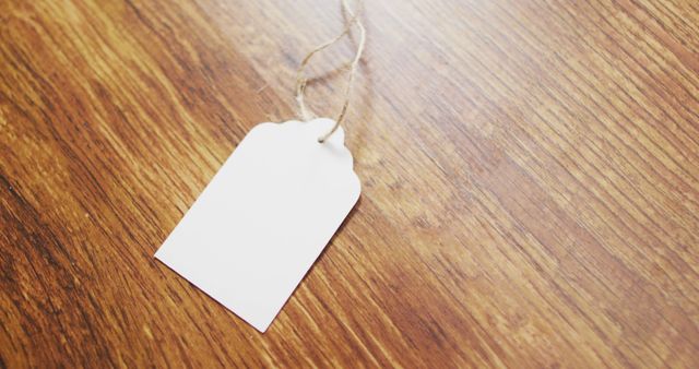 Blank white tag lying on a wooden surface, attached to a piece of string. Ideal for DIY projects, crafting, gift wrapping, product labeling, or event decoration. Versatile for use as a background for design projects requiring space for text or imagery.