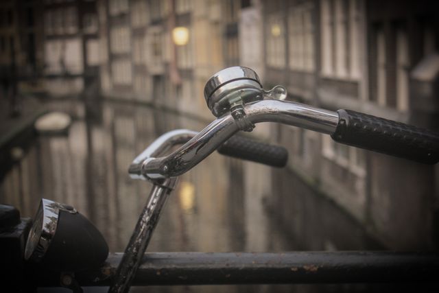 Close-up view of a bicycle handlebar with an urban canal cityscape in the background. Ideal for themes related to cycling, city life, urban exploration, and quaint European settings. Perfect for promotional materials, travel blogs, tourism brochures, and bicycle-themed advertisements.