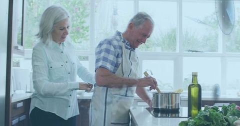 Senior couple cooking together in bright kitchen, man stirring pot as partner ties apron