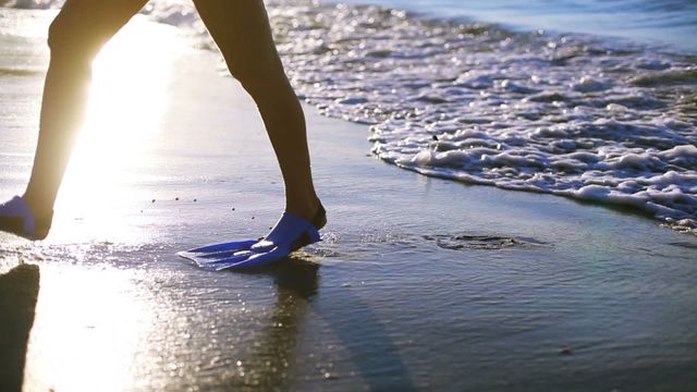 A woman casually walking along a sunny beach wearing swim fins, capturing a serene vacation moment. Perfect for illustrating travel, leisure, relaxation, seaside adventures, summer fun, and outdoor activities. This vibrant scene is ideal for travel brochures, vacation promotions, or beach-themed decorations.