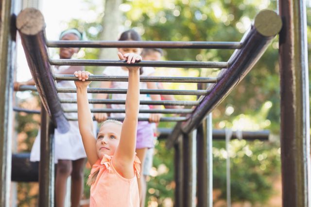 Schoolgirl hanging on monkey bars at playground with friends in background. Ideal for use in educational materials, advertisements for children's activities, fitness programs, and outdoor play promotions. Highlights themes of childhood fun, physical activity, and social interaction.