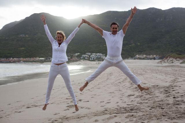 Mature couple enjoying a fun moment by jumping in the air on a sandy beach with mountains in the background. Ideal for use in travel brochures, lifestyle blogs, advertisements promoting active and joyful living, and articles about vacation destinations.