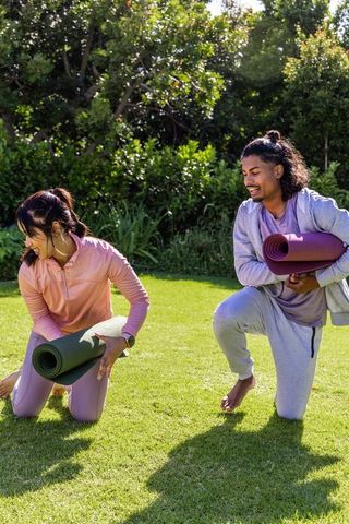 Sunlit garden yoga prep: friends kneeling with rolled mats and smartwatch fitness tracking