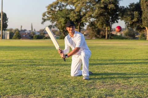 Kneeling cricketer tracking red ball while holding bat on suburban field at golden hour