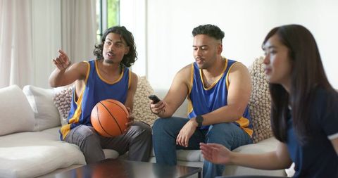 Diverse friends watching basketball game at home wearing blue jerseys holding basketball and remote