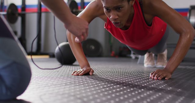 African American woman wearing red tank top doing push-ups on gym floor with a coach motivating her. Suitable for fitness websites, workout inspiration, exercise programs, personal training promotion, and sports equipment ads.