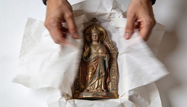 Hands unwrapping antique polychrome wooden madonna holding orb relic on white tissue