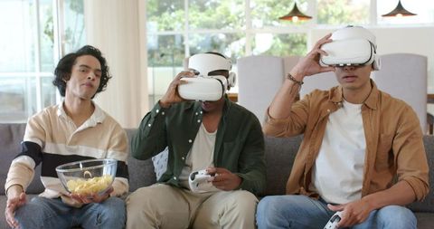 Diverse friends playing VR games on couch wearing white headsets and holding controllers