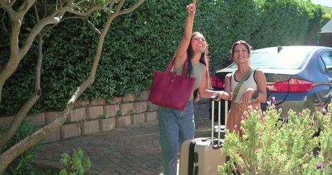 Friends arriving with luggage on sunny driveway pointing upward and smiling