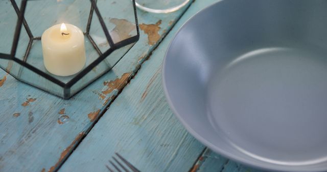 A rustic table setting features a blue plate and a lit candle in a lantern, with copy space. The weathered wooden surface adds a charming, vintage feel to the dining arrangement.