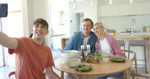 Teen taking selfie with smiling parents at modern kitchen dining table during casual meal
