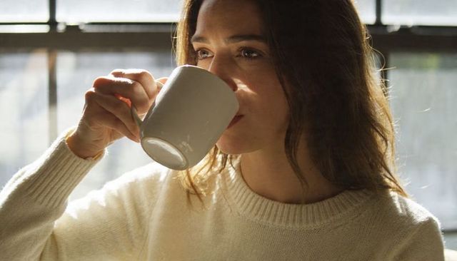 Sunlit woman sipping coffee in cream knit sweater by metal window, serene contemplative
