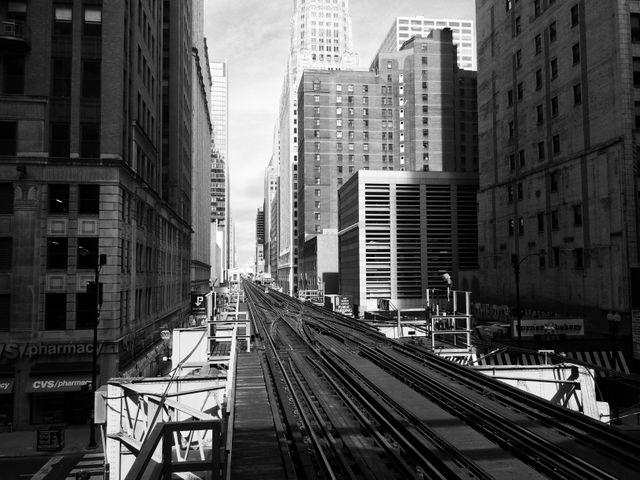 Black and white urban cityscape with elevated train tracks running through downtown. Tall buildings and skyscrapers surround the tracks, creating a dramatic urban scene. Ideal for backgrounds, architectural studies, urban planning projects, transportation-related materials, or retro-themed designs.