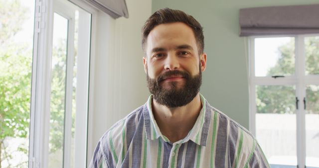 Bearded man smiling near a window at home in casual striped shirt. Ideal for use in lifestyle magazines, advertisements for home-related products, or promotional materials emphasizing relaxed and happy living environments.