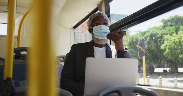 African american senior man wearing face mask talking on smartphone and using laptop while sitting in the bus. hygiene and social distancing during coronavirus covid-19 pandemic.