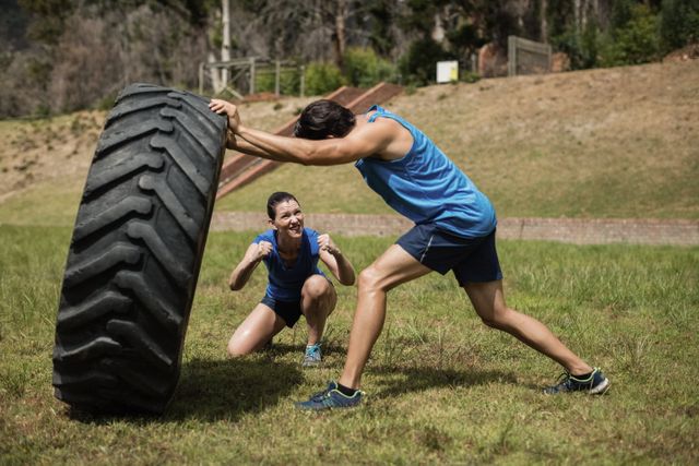 Man flipping a large tire while his trainer cheers him on during an outdoor boot camp. Ideal for use in fitness blogs, workout programs, motivational posters, and advertisements for boot camps or personal training services.