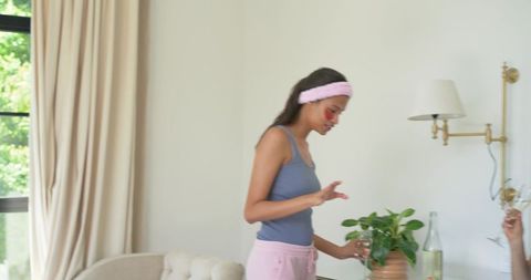 Woman wearing pink headband applying facial patches while tending potted plant in bright living room