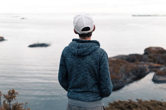 Young man with cap is standing by the serene coastline, looking at the water. The calm sea and rocks in the distance add to the tranquil and peaceful atmosphere. It is ideal for themes of solitude, introspection, leisure, and connection with nature, perfect for use in lifestyle or travel content.