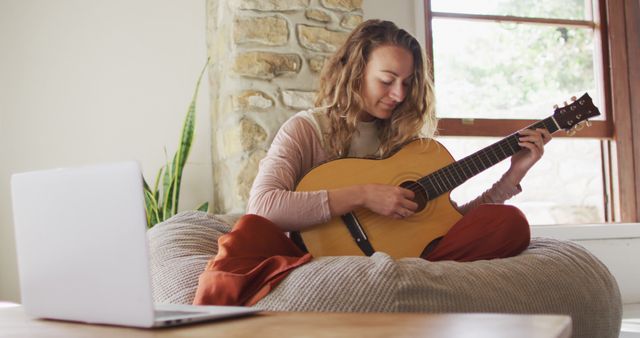 Woman is playing an acoustic guitar while sitting on a comfortable lounge chair, creating a relaxed and cozy home environment. The natural stone wall, plants, and soft seating add to the warm atmosphere. Laptop on nearby table suggests the focus on practice or learning music possibly through online lessons. Suitable for uses related to home living, music lessons online, casual relaxation, hobbies and leisure activities.