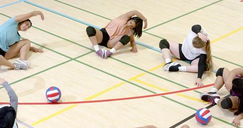 Diverse female volleyball team stretching on indoor court warming up for game, teamwork drills