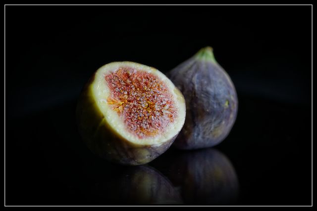 This image shows a close-up of fresh ripe figs, one cut in half, placed on a dark reflective surface. Backlighting highlights the juicy texture and vibrant colors of the figs, adding a gourmand vibe. Perfect for use in food blogs, culinary websites, health and nutrition articles, organic product promotions, and gourmet magazine features.